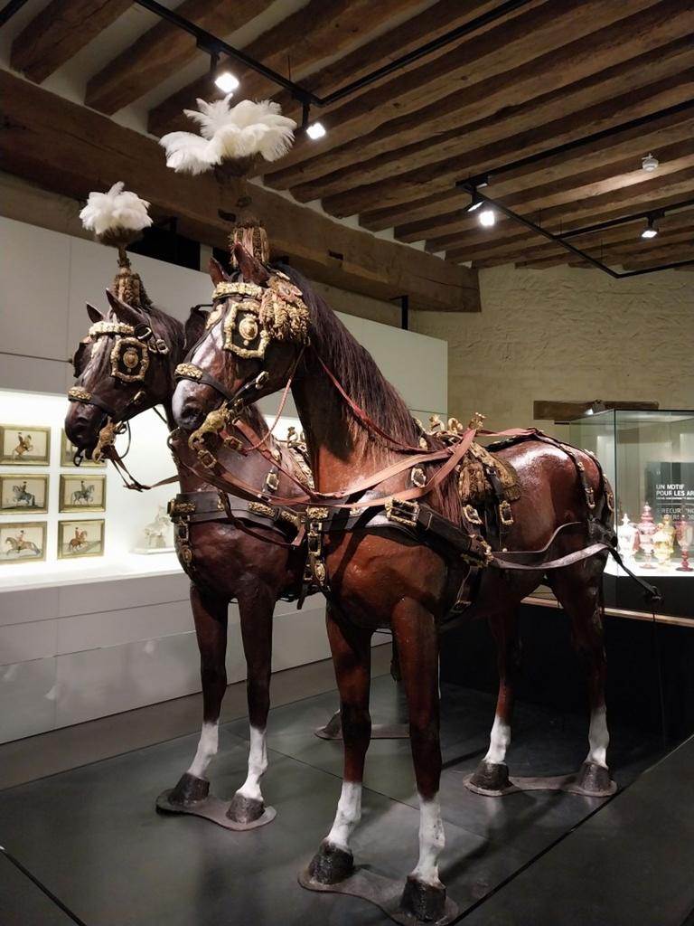 Au premier plan, deux des huit harnais de l’attelage d’apparat commandé par le prince de Condé pour le sacre de Charles X, présentés sur des mannequins de chevaux. On aperçoit à l’arrière-plan une partie des panneaux en Pietra dura et Pietra Paesina attribués à Giuseppe Zocchi (XVIIIe siècle). 