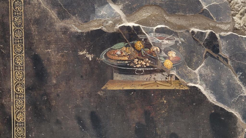 Cette nature morte découverte en 2023 dans l’îlot 10 du Regio IX, à Pompéi, représente un type d’offrande faite aux invités, nommé xenia dans l’Antiquité. Sur un plateau d’argent se trouvent un verre de vin, des fruits secs comme des noix, et une sorte de focaccia.