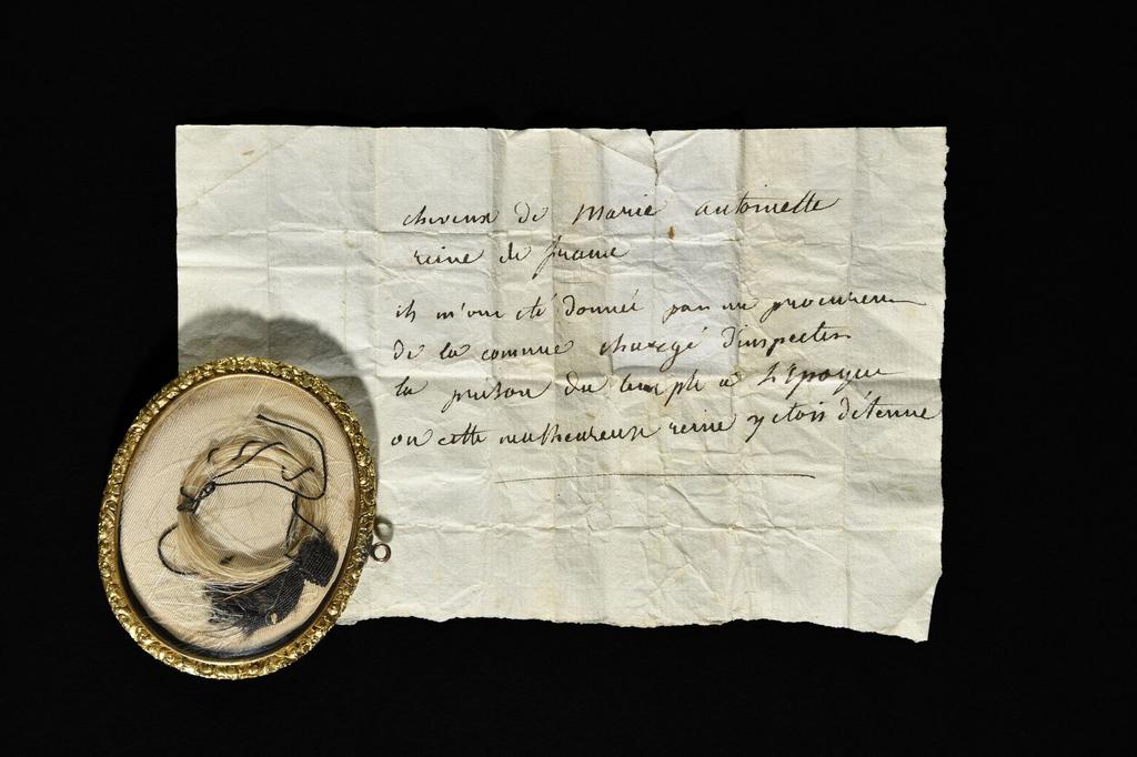 Médaillon contenant une mèche de cheveux blancs de Marie-Antoinette nouée avec un petit ruban et un fil noir. Sous verre, dans un médaillon ovale à suspendre en laiton ciselé à décor de rinceaux et de fleurettes, 6,5 x 5,1 cm.