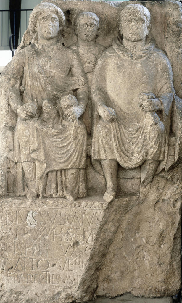 Stèle funéraire d’un couple d’époux de la région de la vallée du Rhin. La femme est habillée à la romaine d’une robe longue (ou stola), par-dessus laquelle elle porte un grand châle rectangulaire (ou palla). À son cou, un gros pendentif est attaché à un collier. L’homme, assis, est vêtu à la gauloise d’un manteau à capuchon. Ier siècle. Mayence, Mittelrheinisches Landesmuseum. 