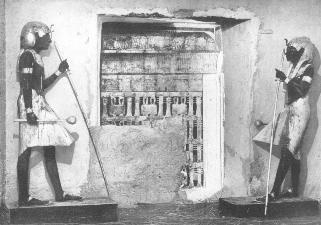 Depuis l’antichambre, deux statues grandeur nature de Toutânkhamon veillent sur la porte donnant vers son sépulcre. Alors que les équipes d’Howard Carter ont, en partie, démonté le mur recouvert des sceaux de la Nécropole, on distingue à travers l’ouverture la première des chapelles. Photographie d’Harry Burton prise au moment de l’ouverture de la chambre funéraire, le 16 février 1925.