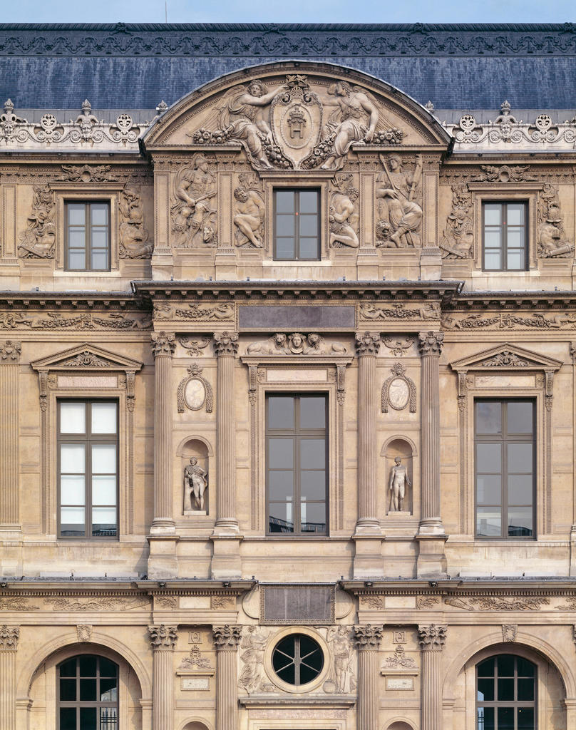 Détail du décor de l’attique de l’avant-corps sur la façade Henri II, sculpté par Jean Goujon entre1546 et 1553 : en haut, deux Victoires encadrent les armes royales, surplombant Mars, Bellone et deux captifs enchaînés ; courant au-dessus des fenêtres de l’étage, les frises d’enfants ; au-dessus de la porte, l’oculus encadré de figures.